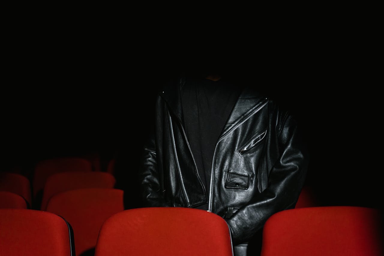 about-us Individual wearing black leather jacket sits among red seats in a dimly lit cinema.